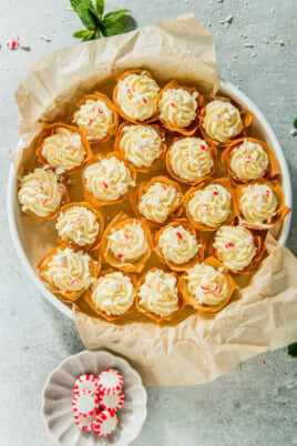 A plate of cream-topped tarts with red and white sprinkles, placed on parchment paper. A small dish with peppermint candies is nearby, on a light-colored surface.