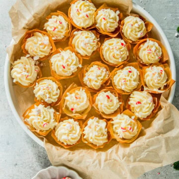 A plate of cream-topped tarts with red and white sprinkles, placed on parchment paper. A small dish with peppermint candies is nearby, on a light-colored surface.