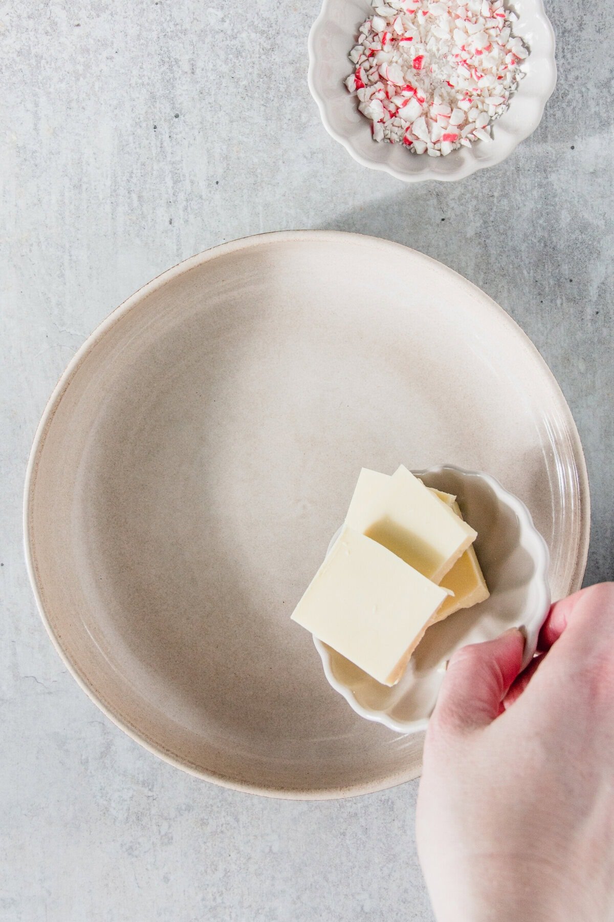 Place a small plate of butter on a large empty plate by hand. In the background, on a light gray surface, is a bowl of crushed peppermint candy.