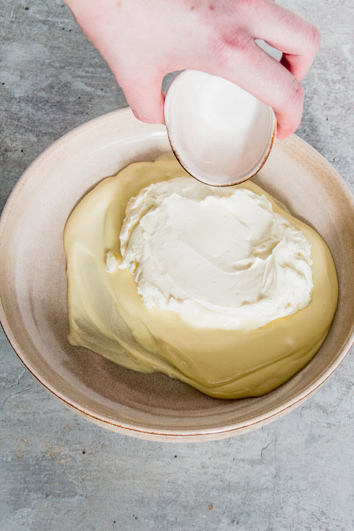 A hand pours cream from a small bowl into a larger bowl filled with cream on a gray surface.