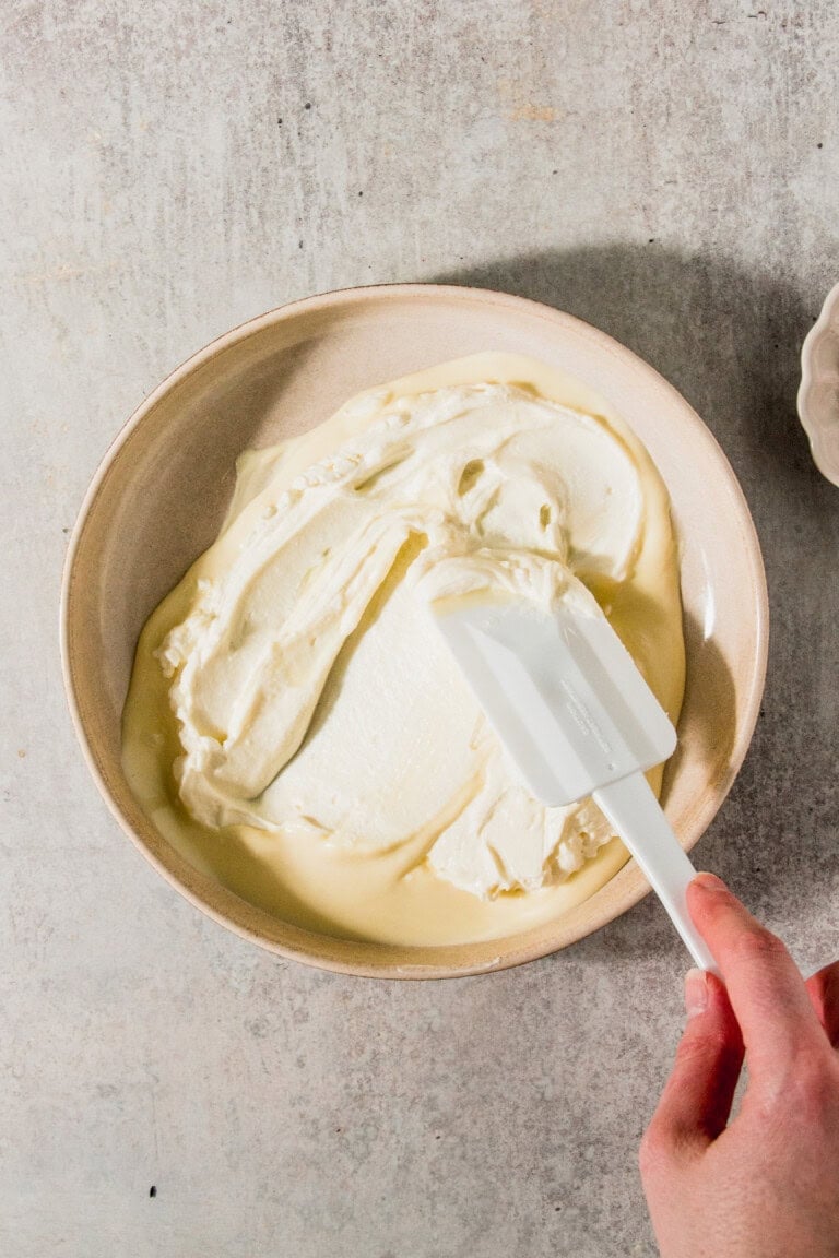 A hand using a white spatula to mix creamy batter in a beige bowl on a light gray surface.