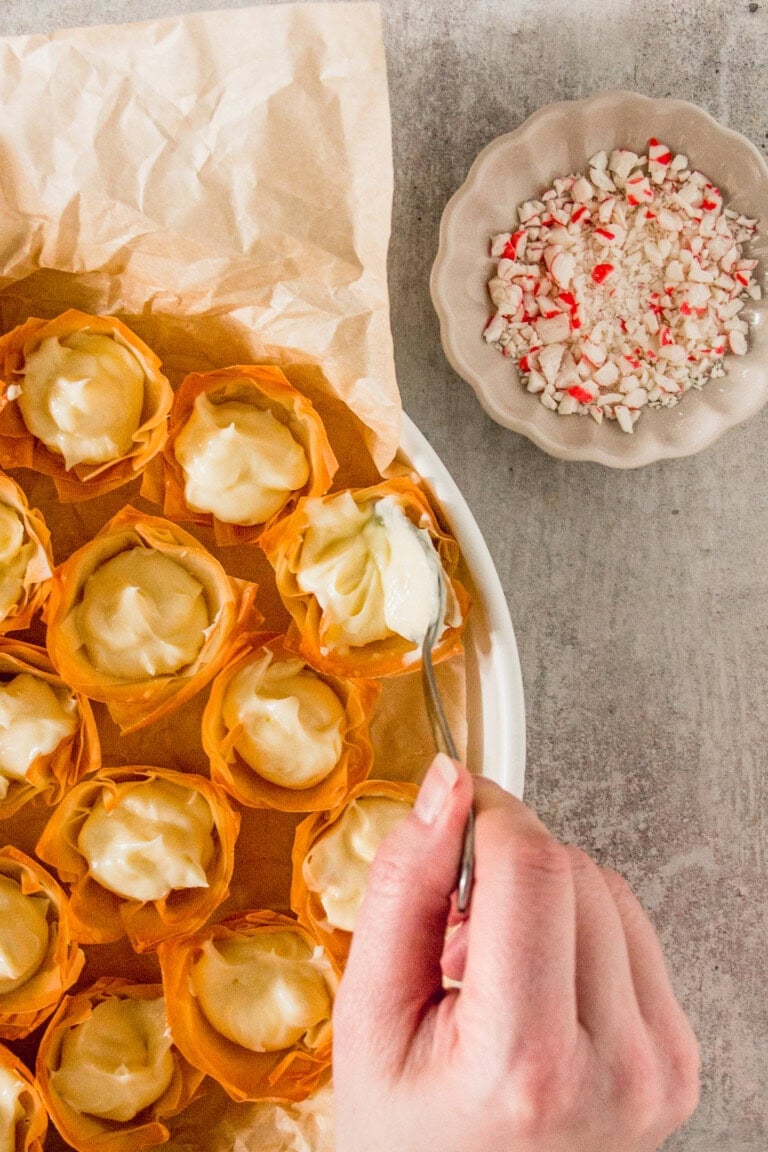 A hand is filling phyllo dough cups with a cream mixture. A bowl of crushed peppermint candy is nearby.