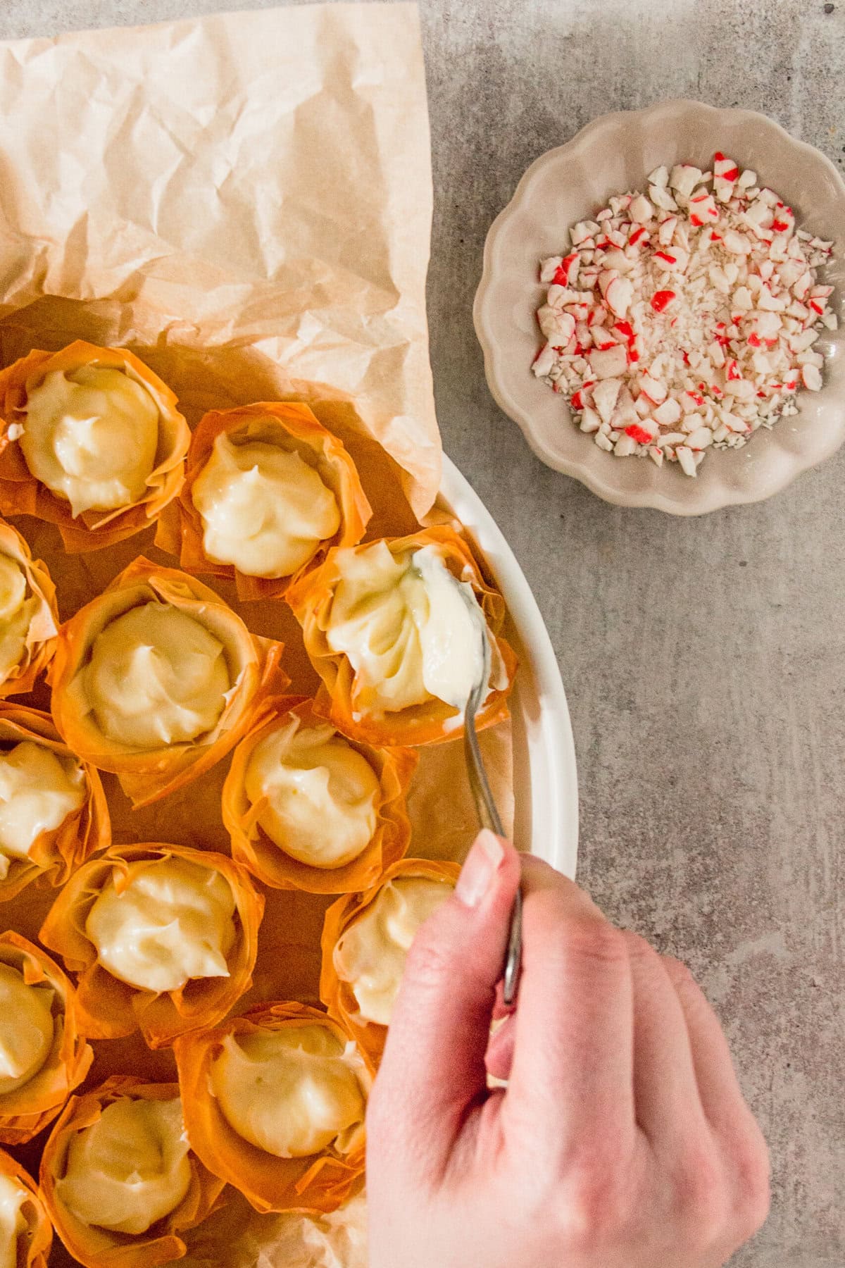 A hand fills phyllo dough cups with a cream mixture. Nearby is a bowl of crushed peppermint candy.