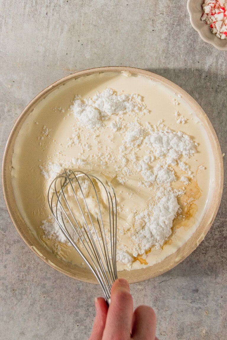 A hand whisking ingredients in a beige bowl on a textured surface.