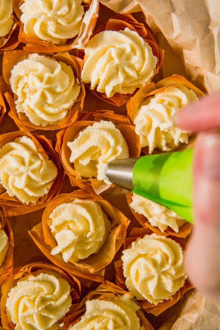 A hand uses a piping bag to add cream to several cupcakes with orange wrappers on a parchment paper background.