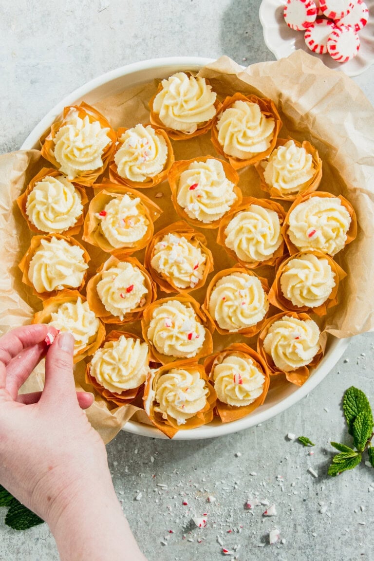 A hand is garnishing a tray of mini white frosted cupcakes with crushed candy canes.