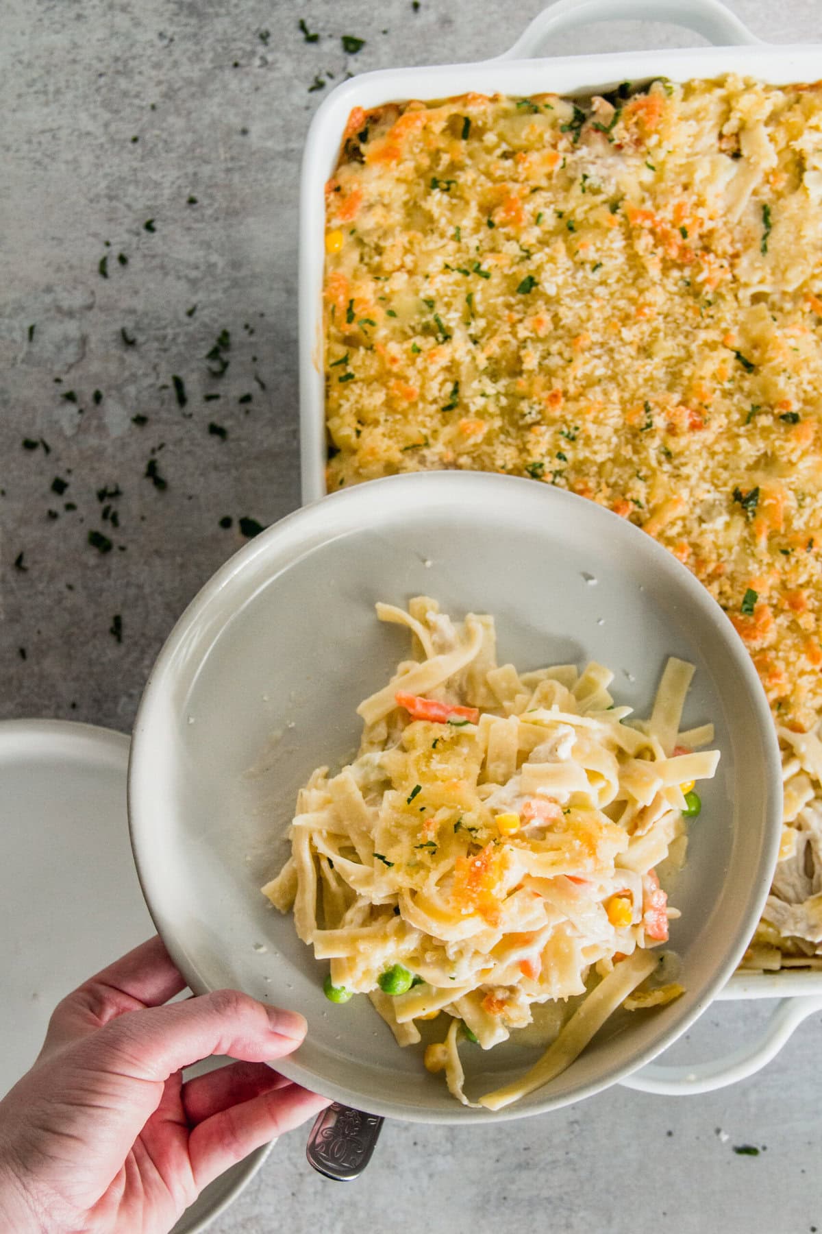 A hand serves a portion of chicken casserole with pasta and vegetables from a white baking dish onto a plate. The casserole, featuring a breadcrumb topping, is beautifully sprinkled with chopped herbs.