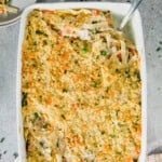 A savory chicken casserole topped with breadcrumbs in a white dish, revealing noodles, peas, and carrots. A serving is plated in the background.