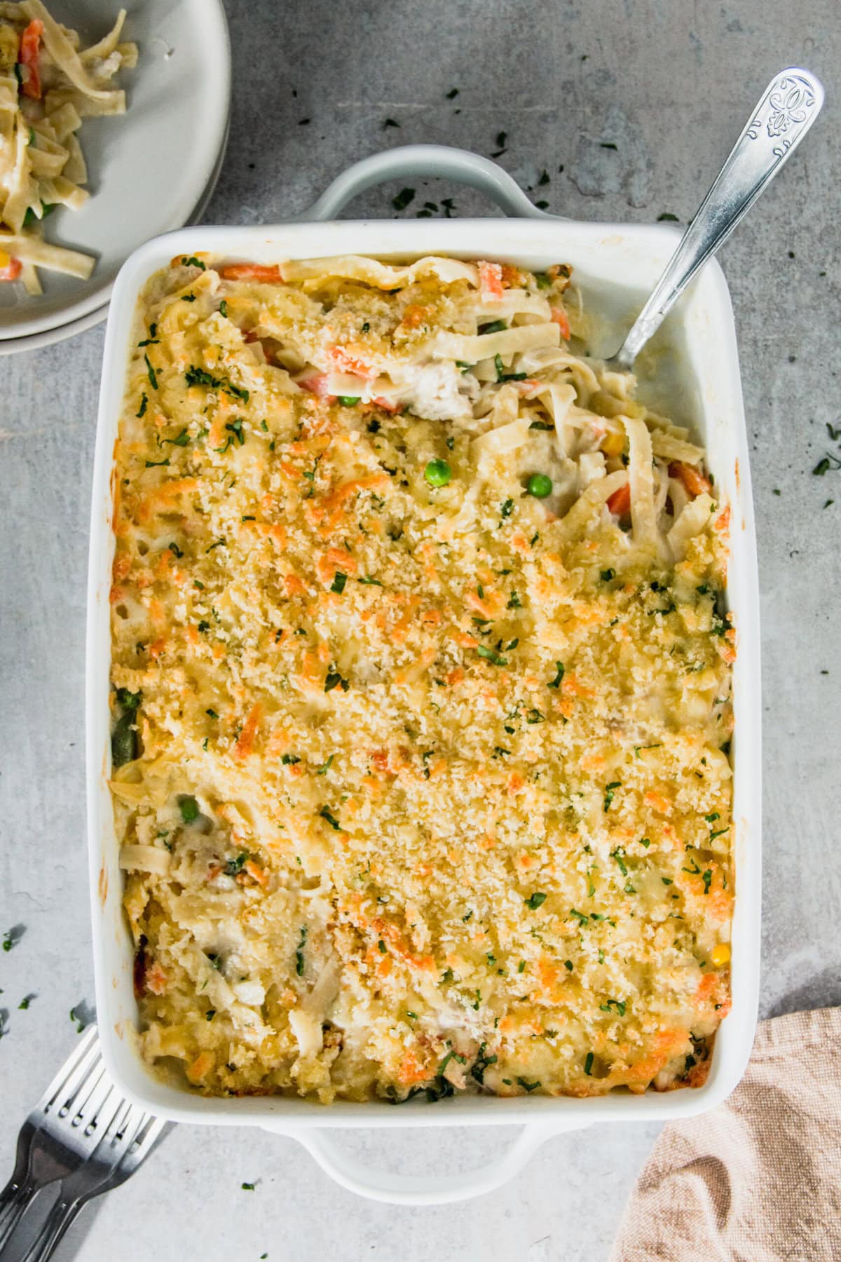 A savory chicken casserole topped with breadcrumbs in a white dish, revealing noodles, peas, and carrots. A serving is plated in the background.