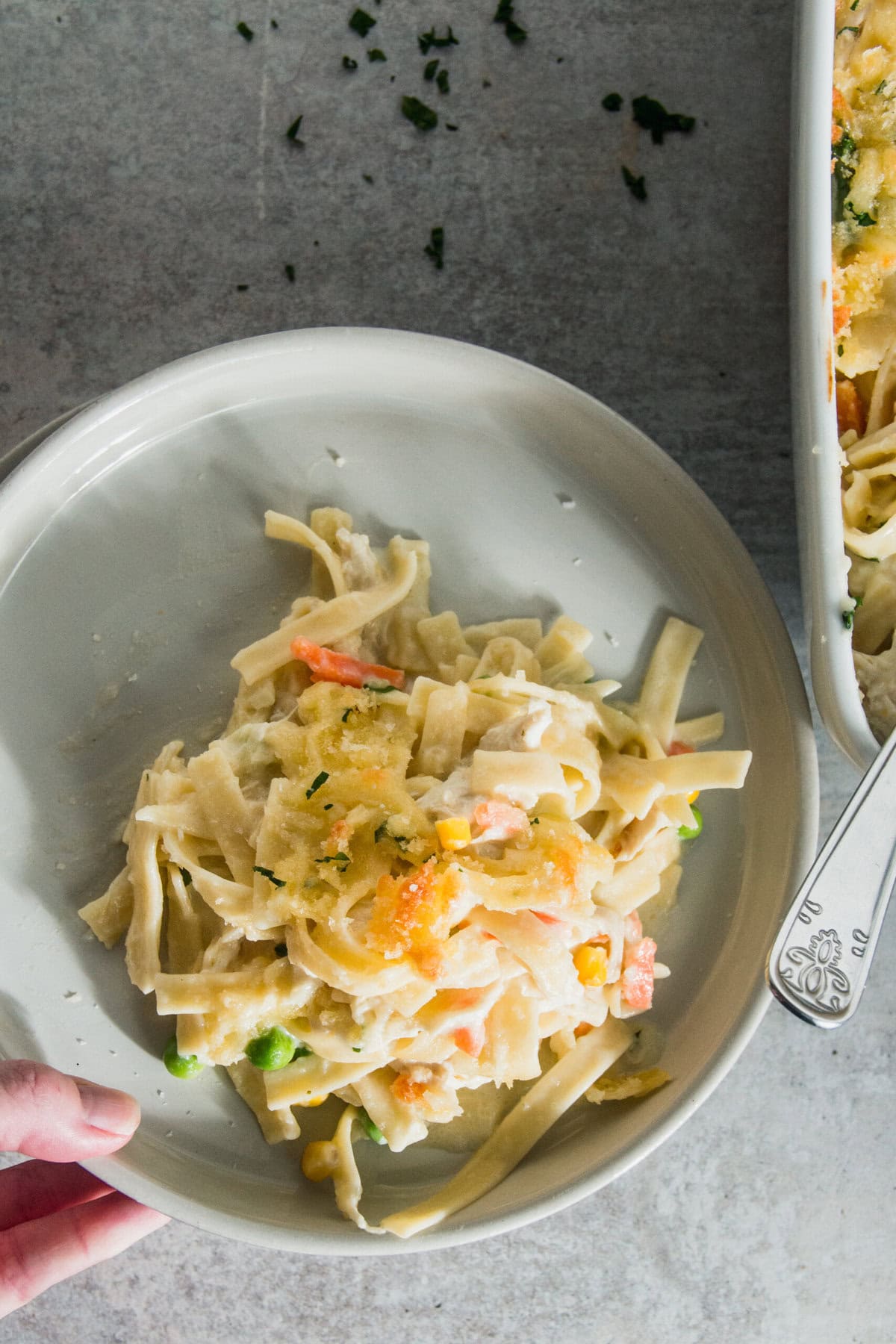 A hand holds a plate of creamy pasta with vegetables next to a delectable chicken casserole dish on a gray surface.