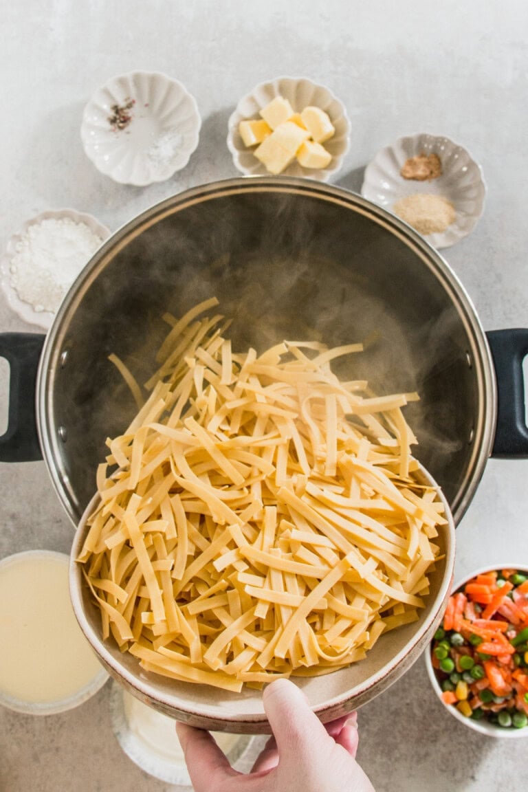 A hand pours uncooked pasta strips into a large pot. Various ingredients in small bowls surround the pot, including spices, butter, and vegetables. Steam is rising from the pot.