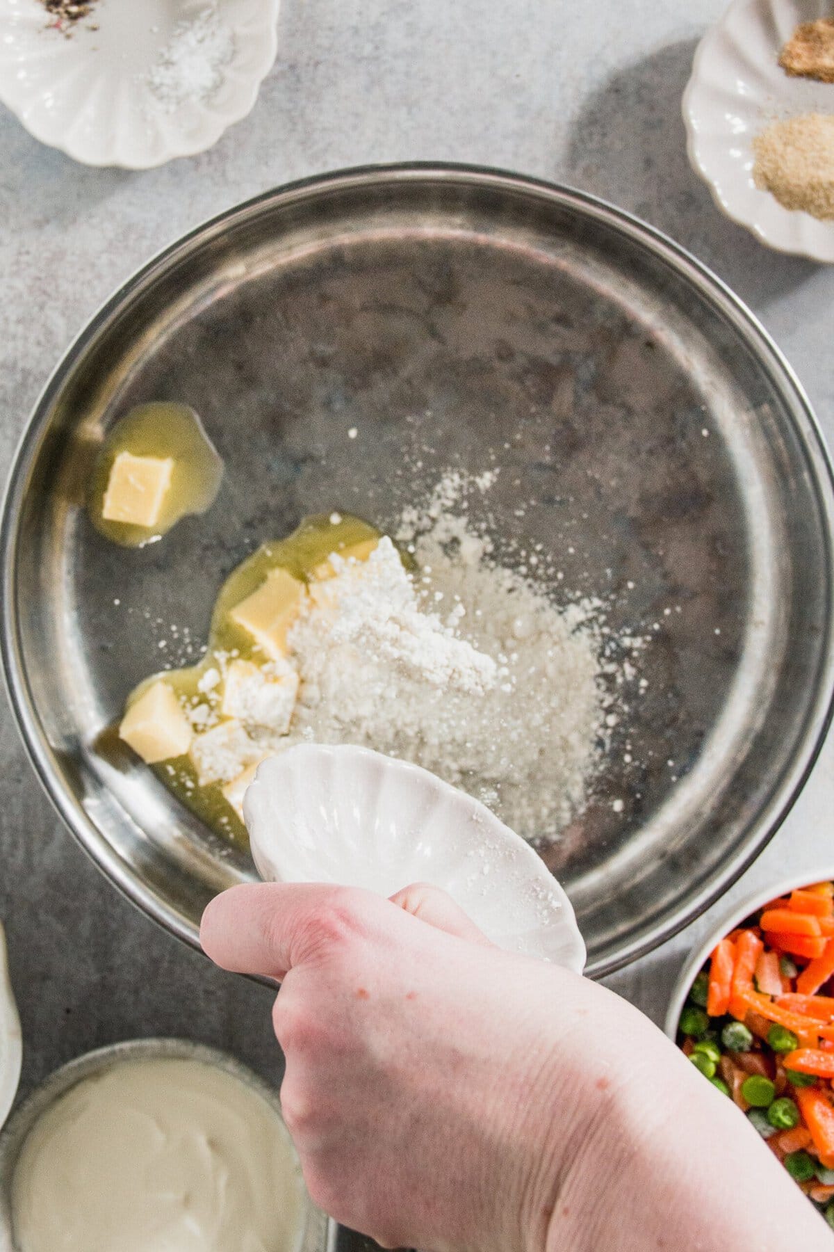 Hand adding flour to a bowl with butter and sugar, surrounded by small plates and bowls of ingredients, including chopped vegetables.