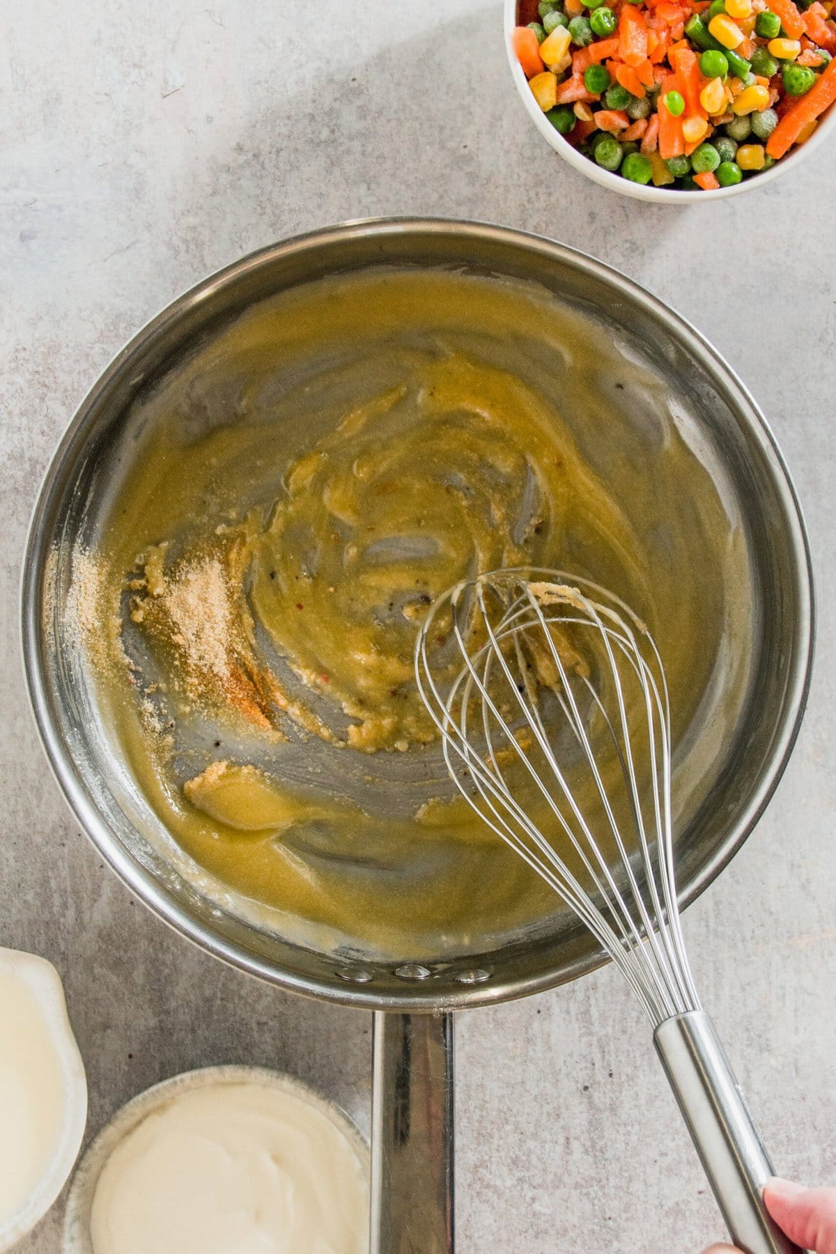 A person is whisking a brown sauce in a pan. Nearby are bowls of cream, mixed vegetables, and a light liquid.