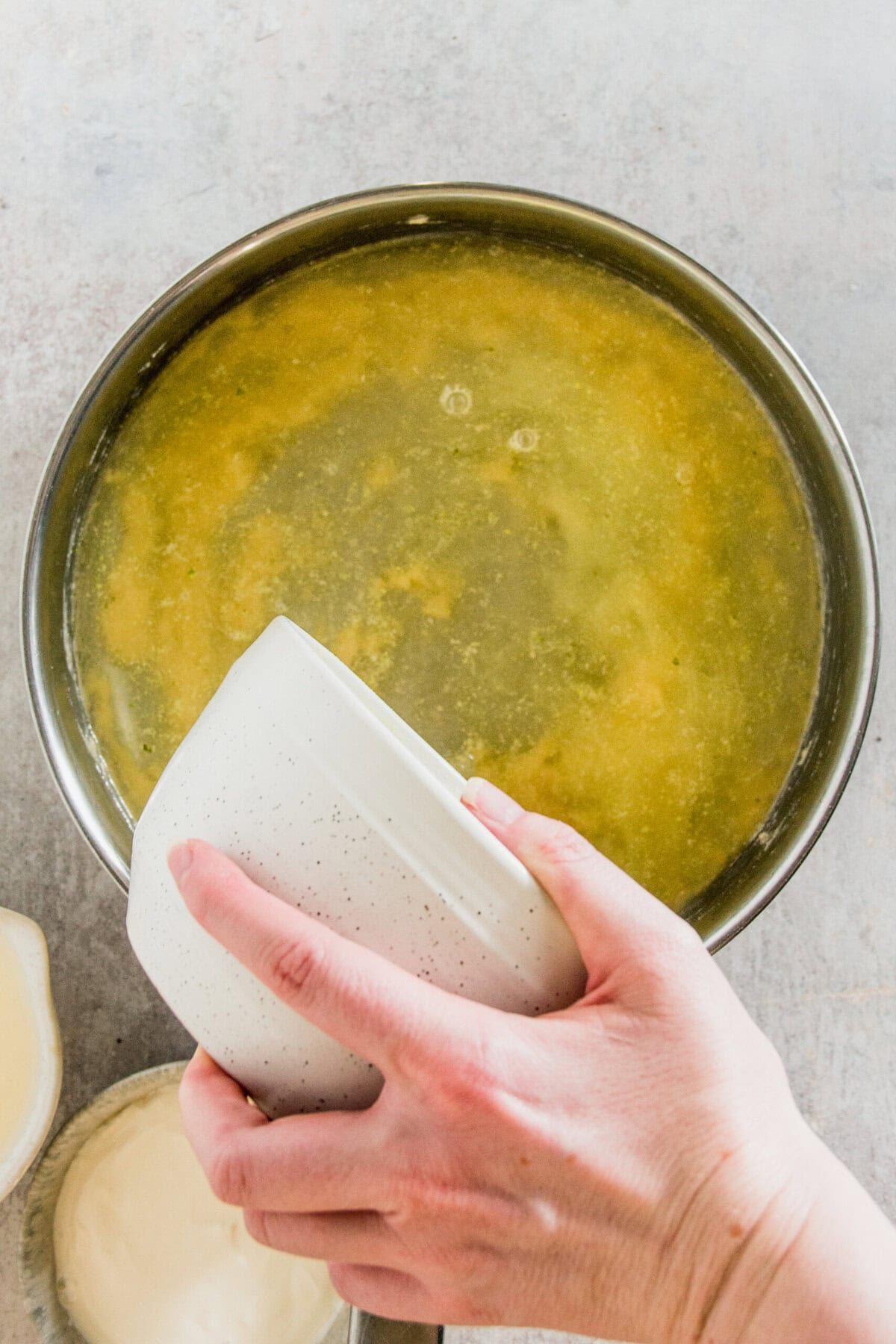 Hands pouring liquid into a bowl containing yellow broth or soup.