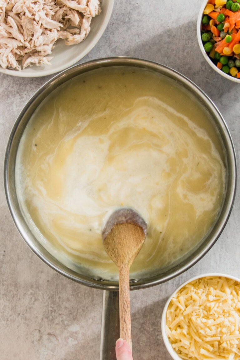 A pot of creamy soup being stirred with a wooden spoon. Shredded chicken, mixed vegetables, and grated cheese are in bowls nearby.