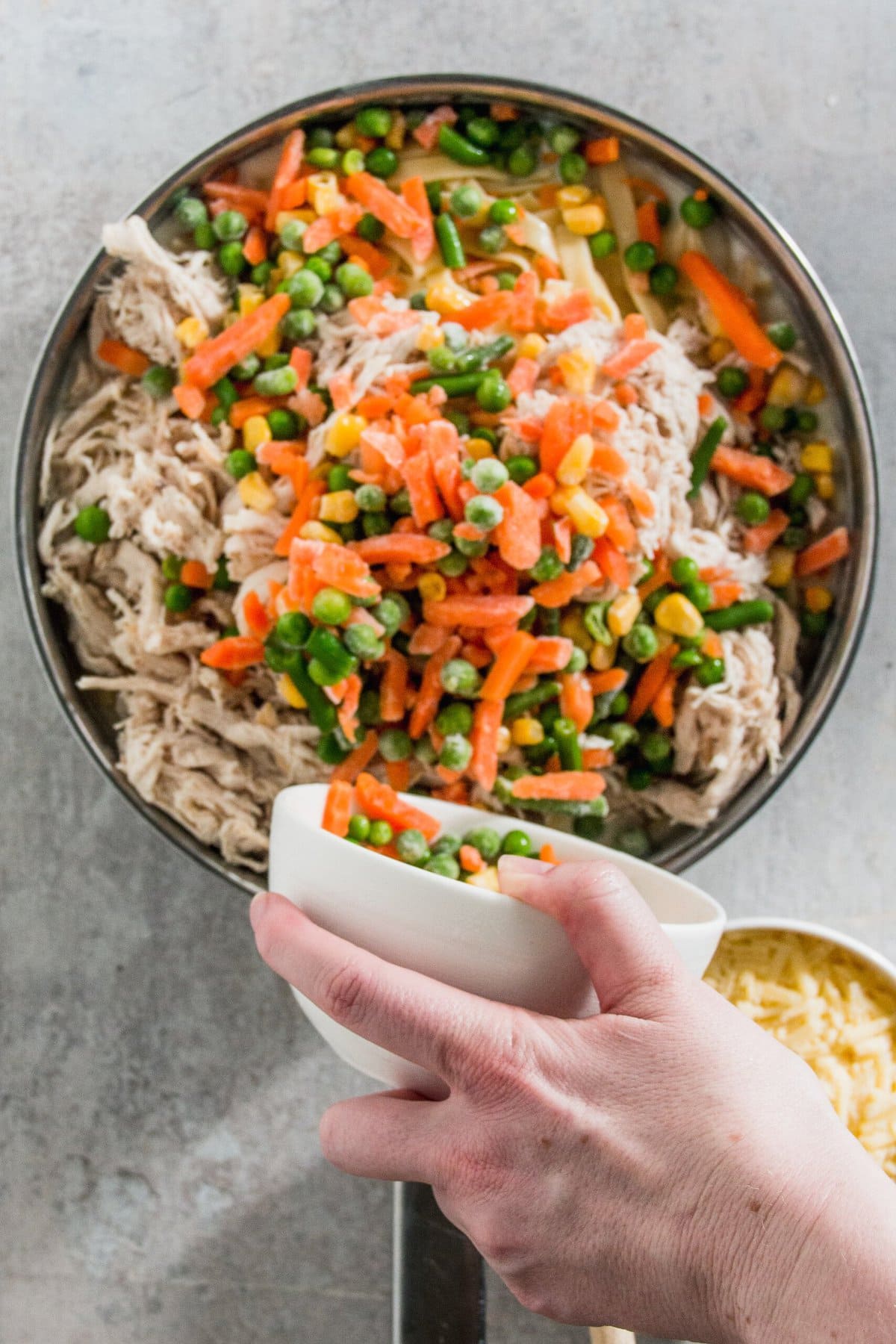 A hand holding a small white bowl hovers above a larger bowl filled with shredded chicken and mixed vegetables, including carrots, peas, and corn.