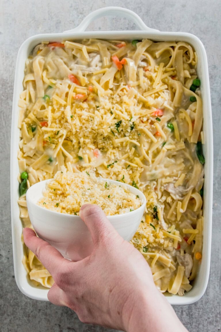 A hand holds a bowl of breadcrumbs above a baking dish filled with pasta, vegetables, and cheese.