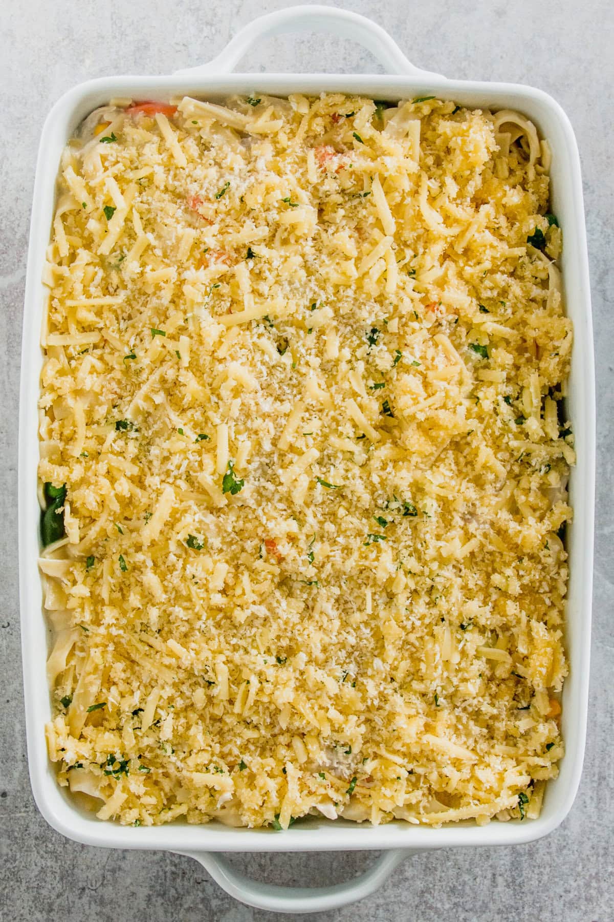 A rectangular baking dish filled with unbaked pasta casserole topped with grated cheese and breadcrumbs.