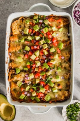 A white dish filled with enchiladas, topped with diced avocado, tomato, red onion, and cilantro. Bowls of sour cream, red onions, and cilantro are beside the dish.
