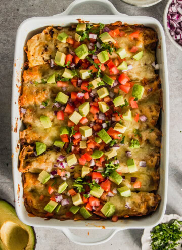 A white dish filled with enchiladas, topped with diced avocado, tomato, red onion, and cilantro. Bowls of sour cream, red onions, and cilantro are beside the dish.