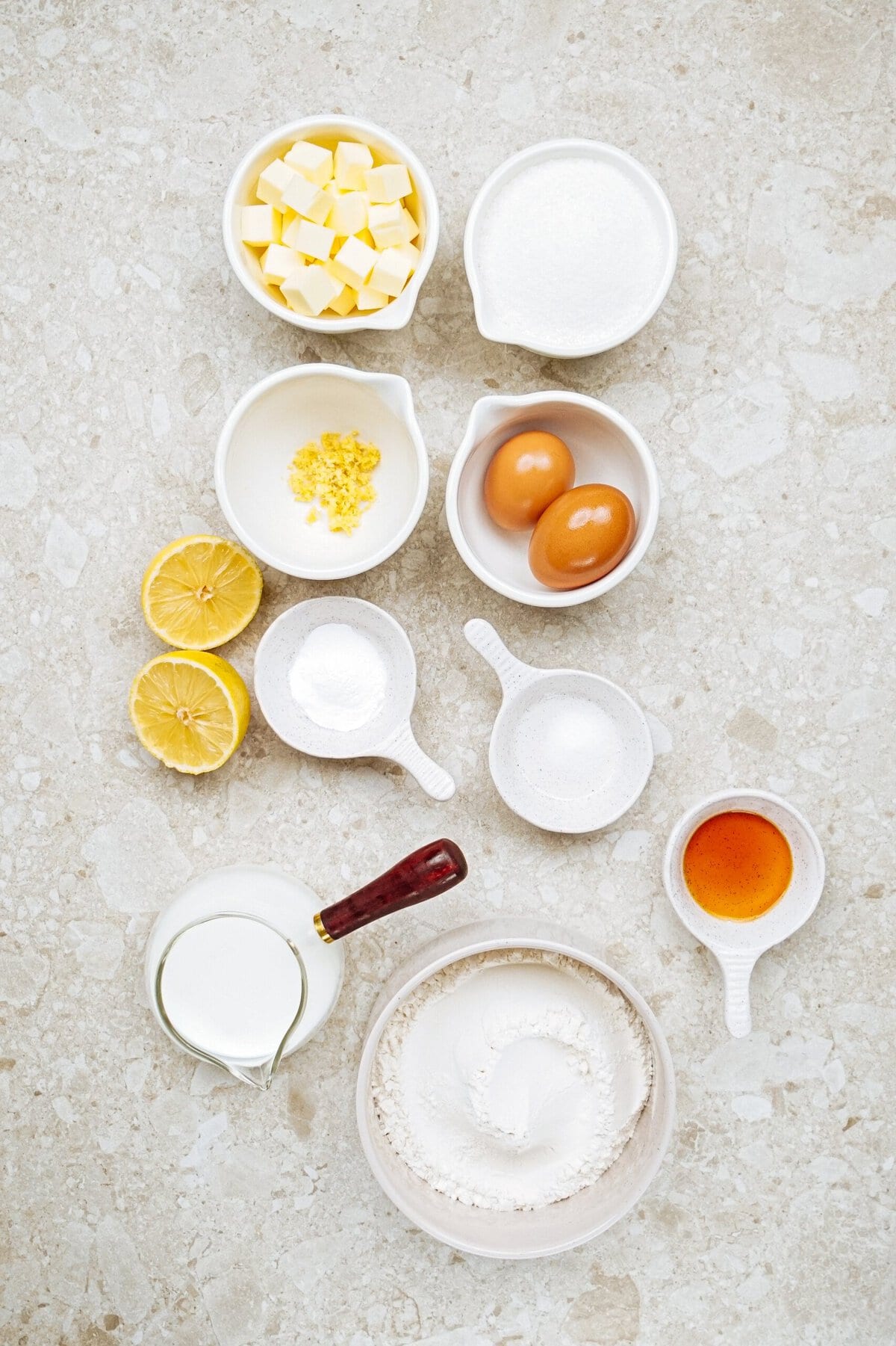 Ingredients for baking arranged on a countertop include butter cubes, sugar, lemon zest, eggs, lemon halves, baking powder, salt, milk, flour, and vanilla extract.