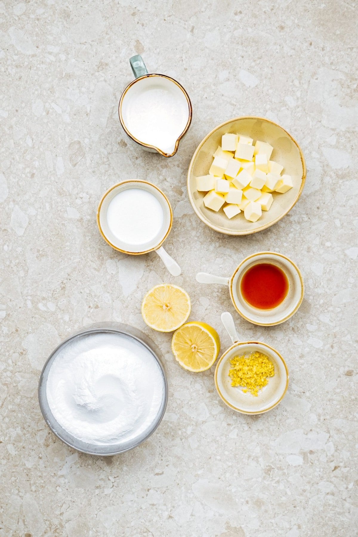 Various baking ingredients including whipped cream, milk, butter cubes, vanilla, lemon slices, and lemon zest arranged on a light-colored surface.