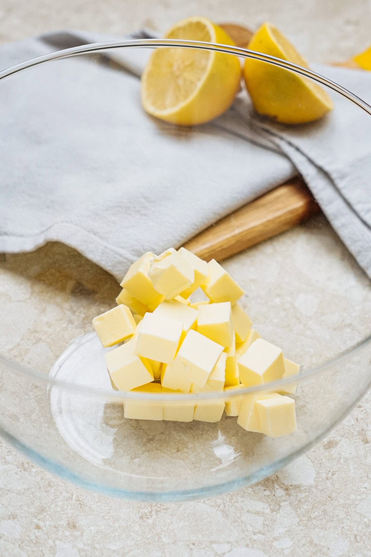 Diced butter in a glass bowl on a countertop, with halved lemons and a gray cloth nearby.