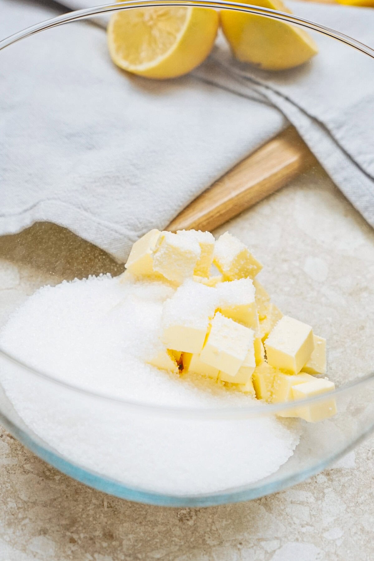 A glass bowl contains sugar and diced butter on a counter. A lemon half and a gray cloth are in the background.