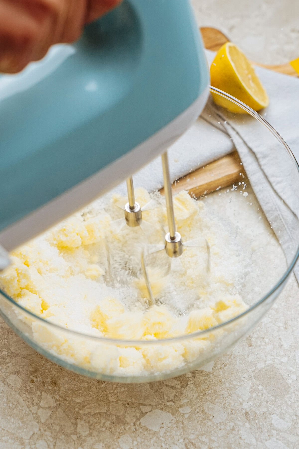 A hand mixer blending butter and sugar in a clear glass bowl, with a halved lemon and a towel visible in the background.