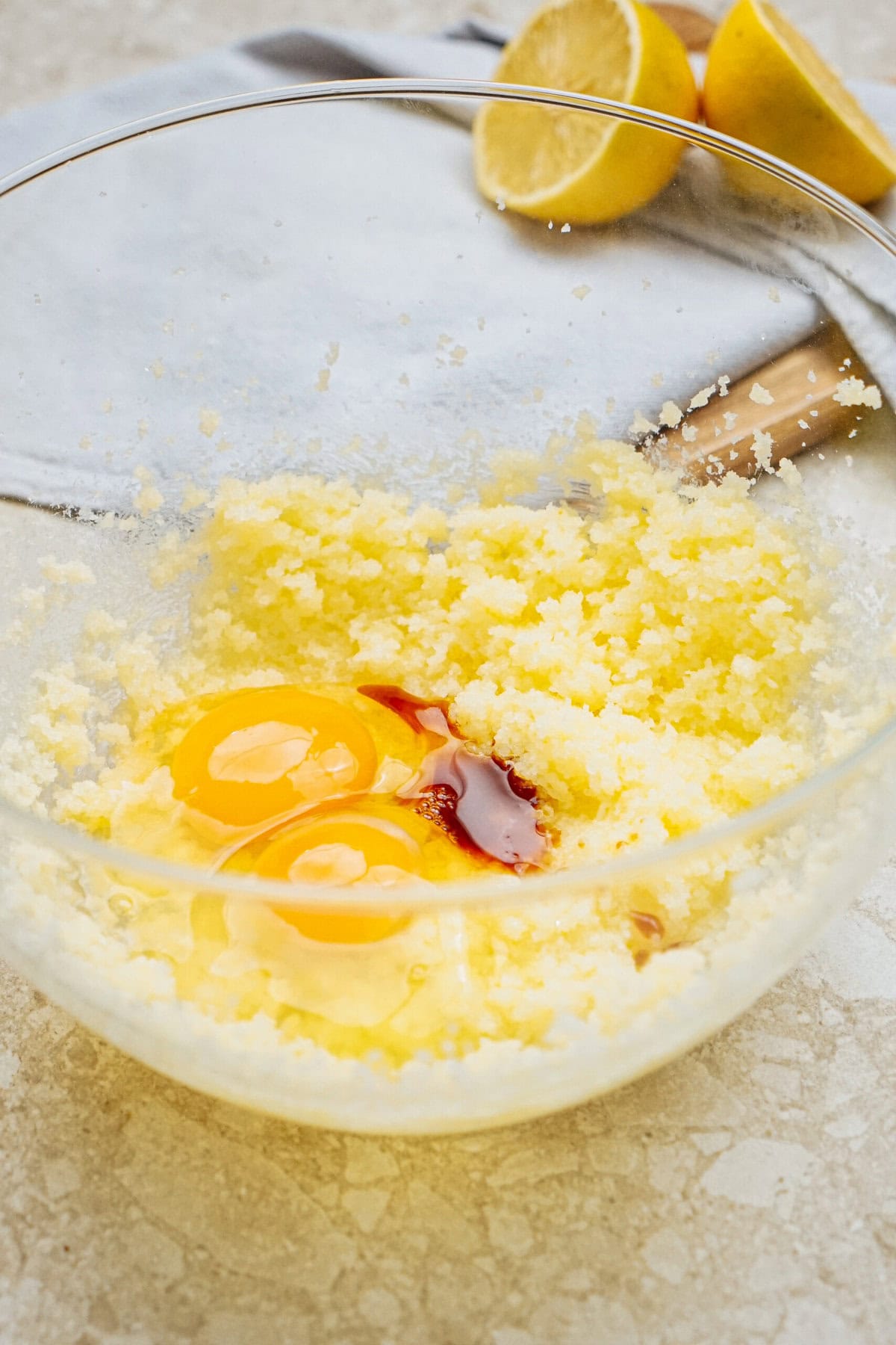 A glass bowl with creamed butter and sugar, two cracked eggs, and vanilla extract on a marble counter. Halved lemons and a kitchen towel are in the background.