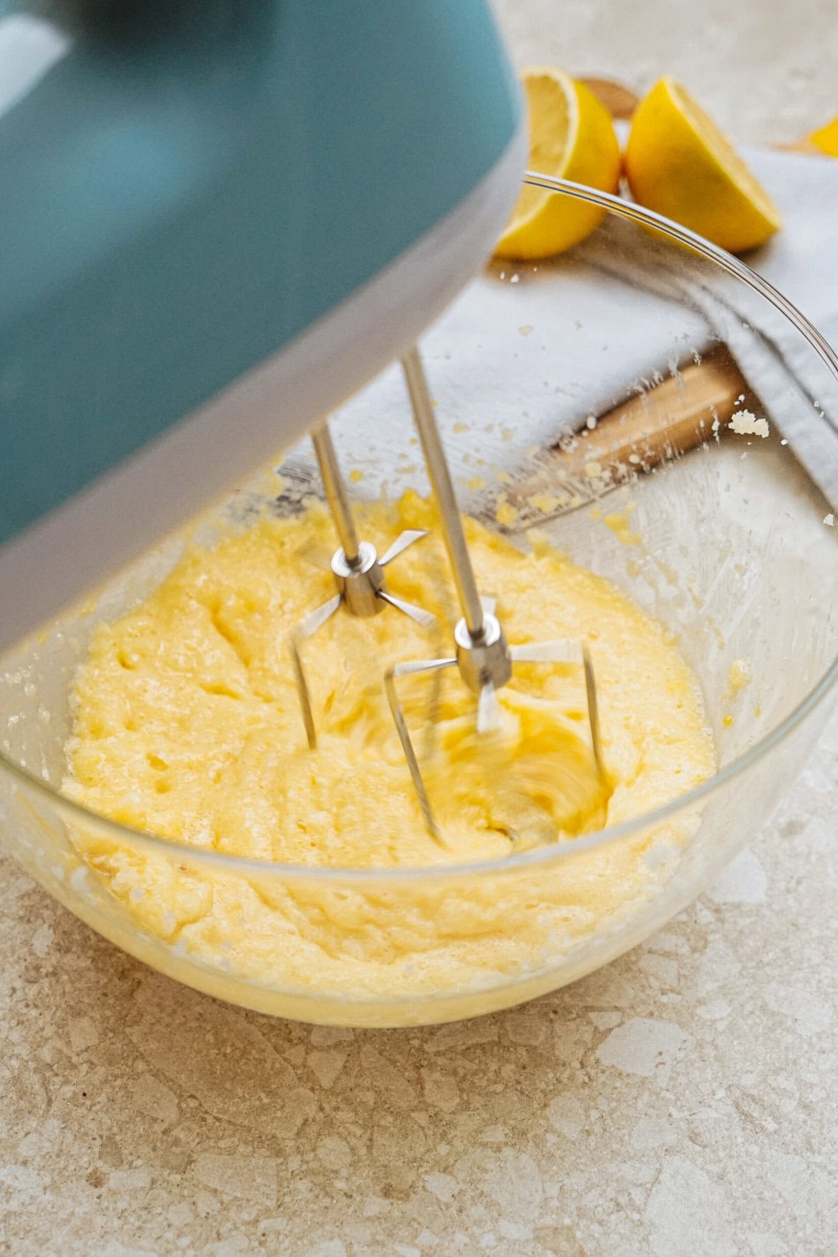 A mixing bowl with yellow batter being mixed by an electric hand mixer. Halved lemons and a white cloth are in the background on a counter.