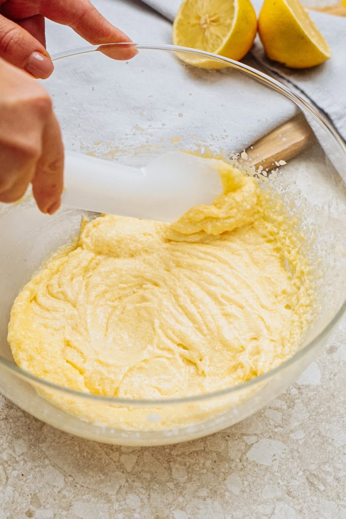 A person mixing a creamy batter in a glass bowl with a spatula. Halved lemons are visible in the background.