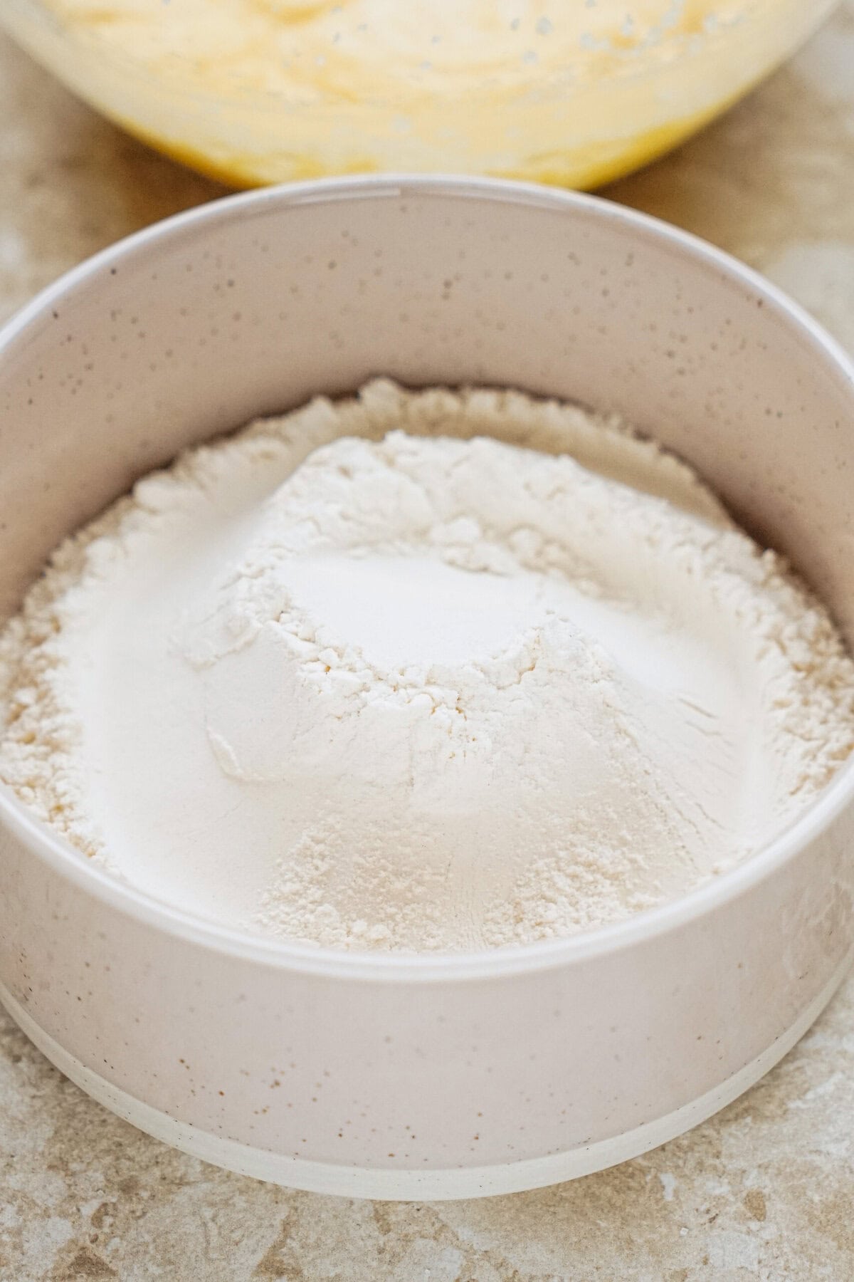 A bowl of white flour on a countertop, with a yellow mixture visible in the background.