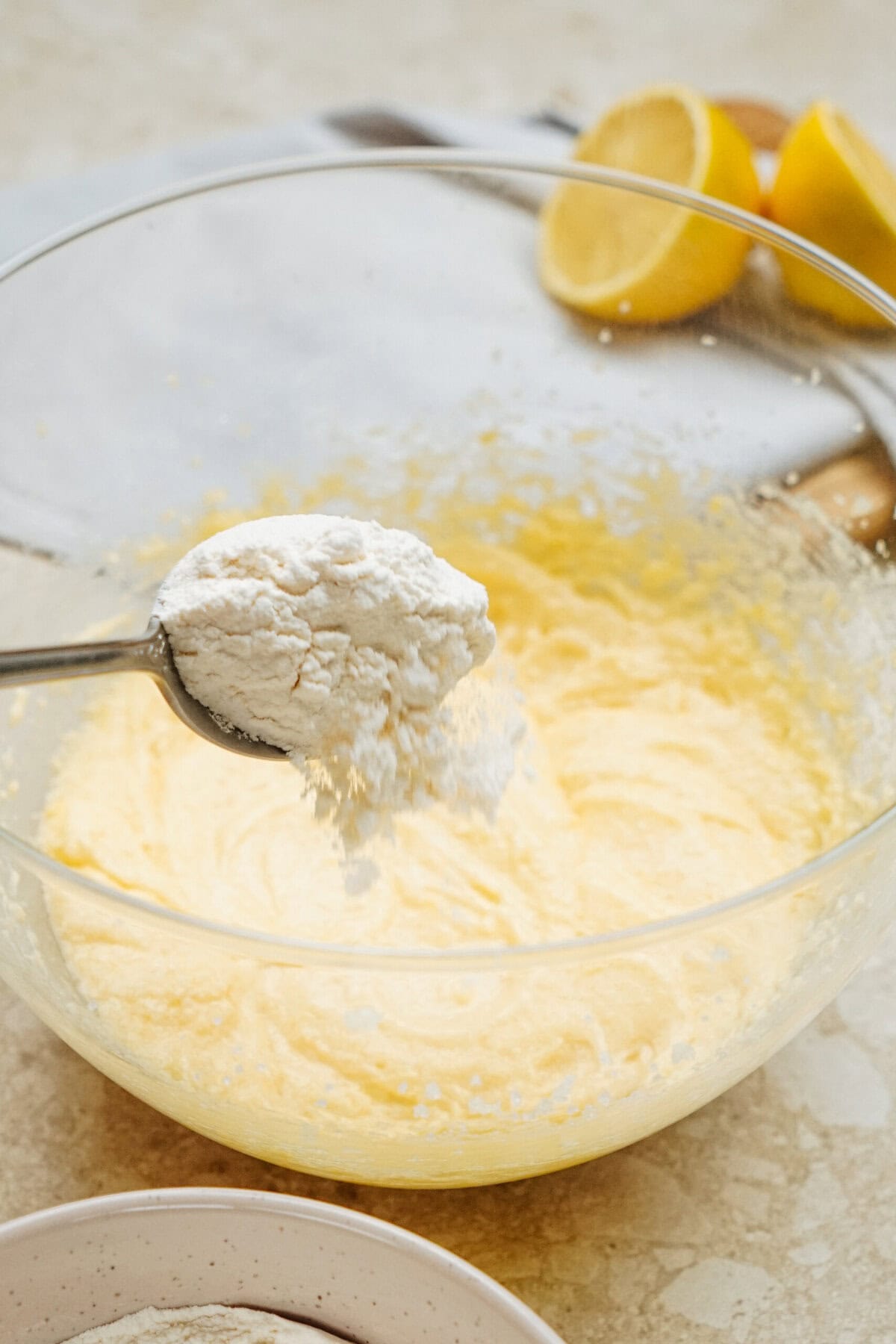 A glass bowl with yellow batter, a spoon holds flour over it. Halved lemons lie in the background.