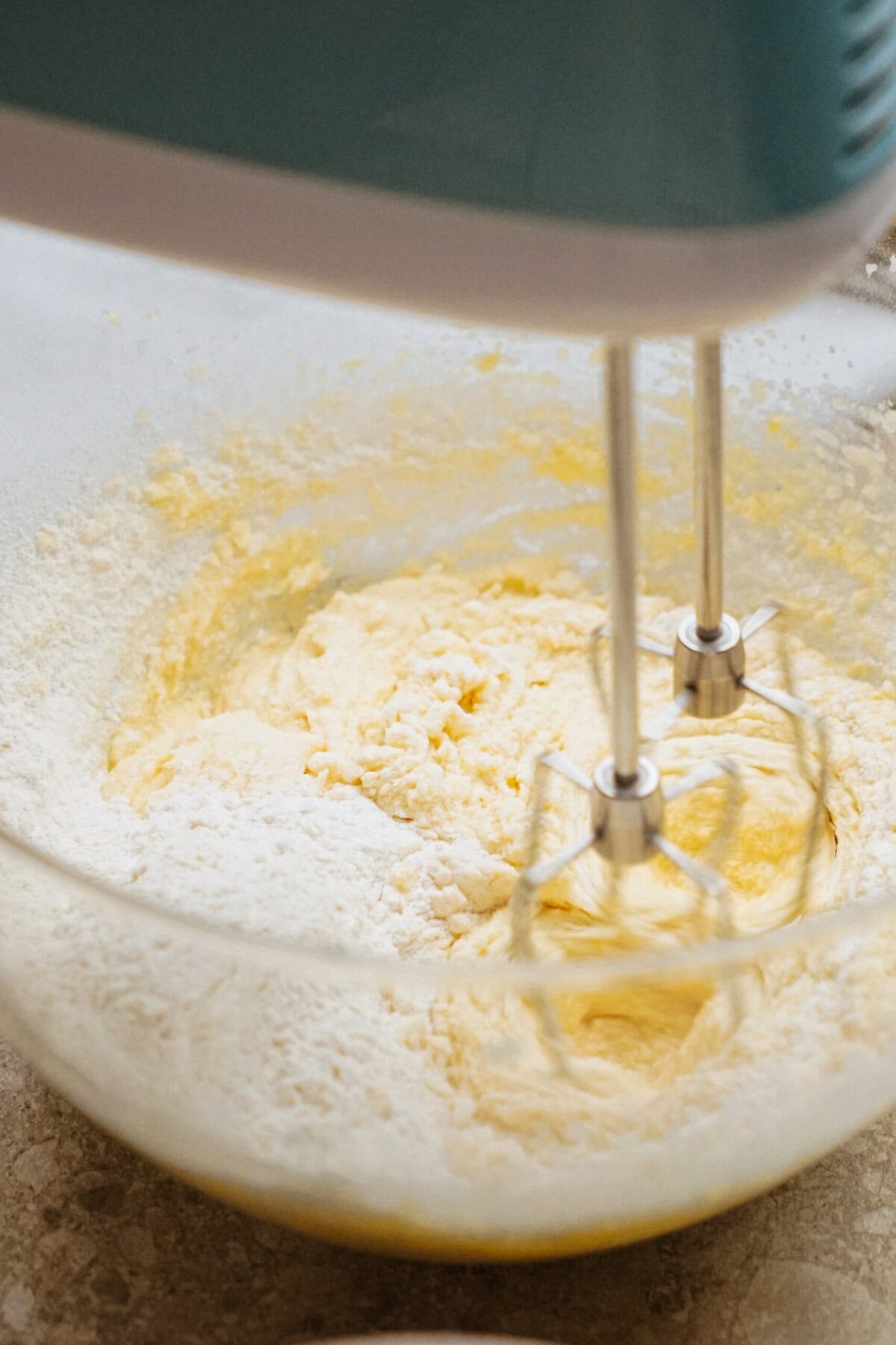 A hand mixer blending flour into a creamy batter in a clear glass bowl.