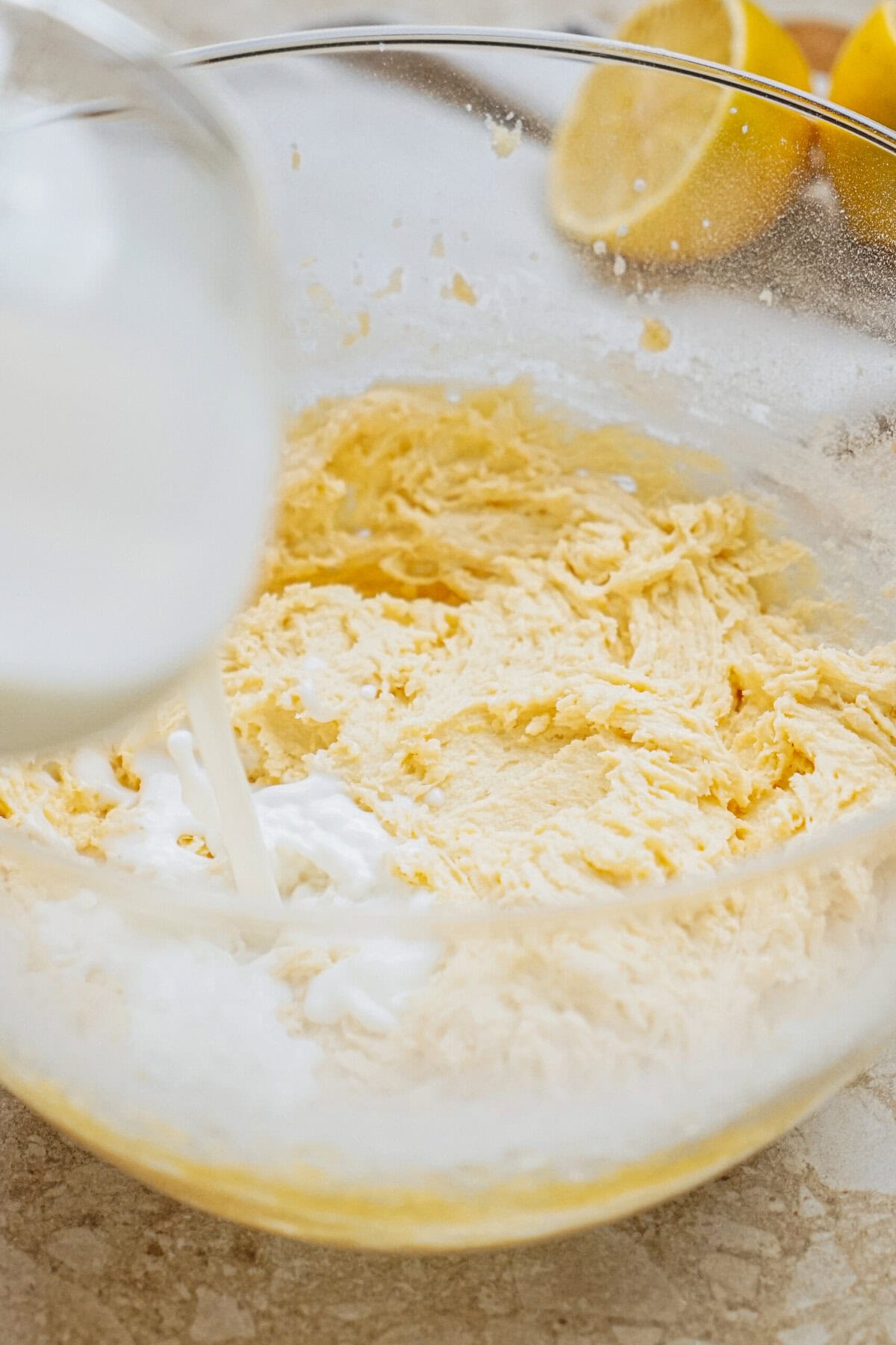 A glass bowl with cake batter is being poured with milk, with halved lemons in the background.