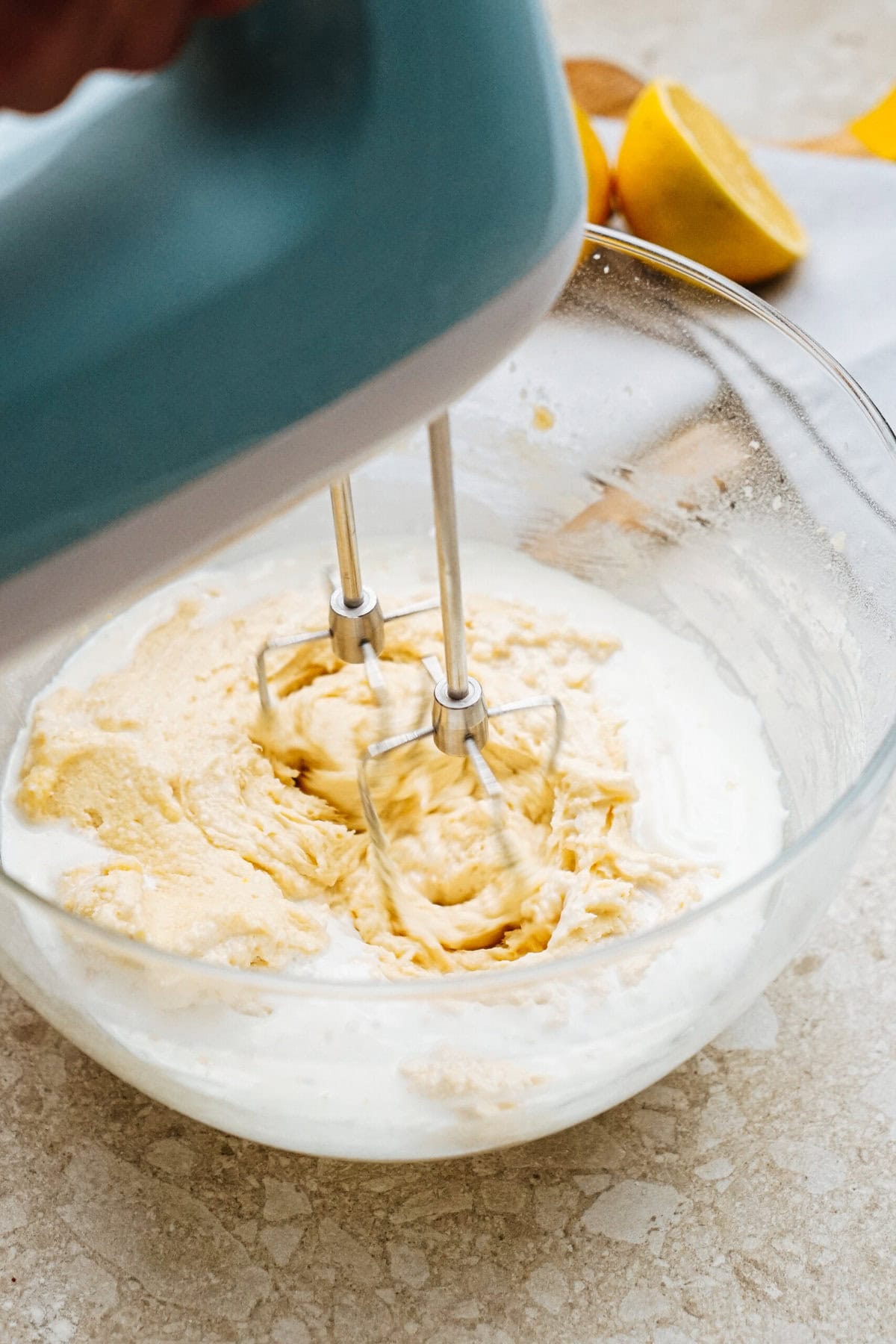 A handheld electric mixer beating creamy yellow batter in a glass bowl on a speckled countertop.