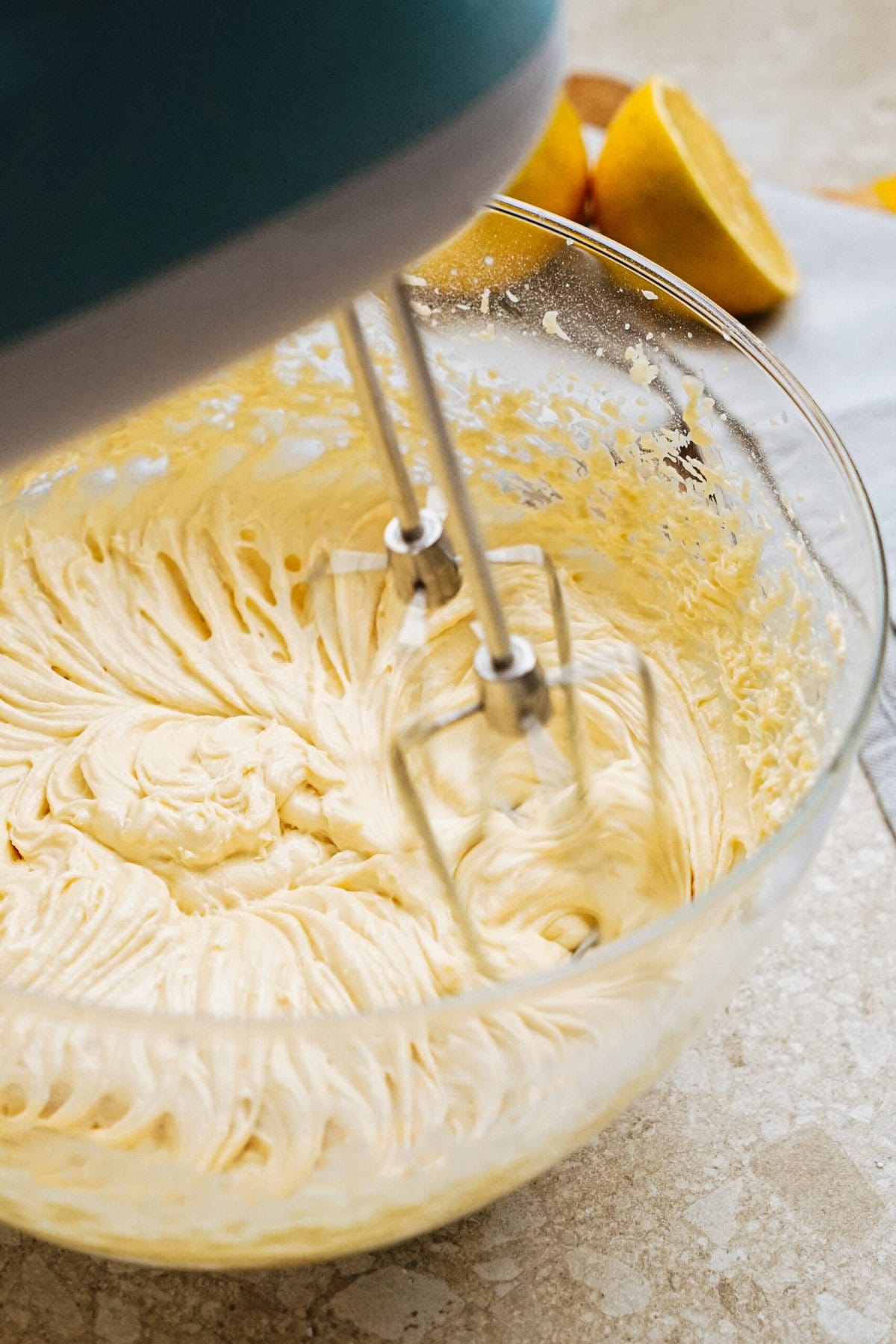 A hand mixer is blending batter in a glass bowl on a kitchen counter. Two halved lemons are in the background.