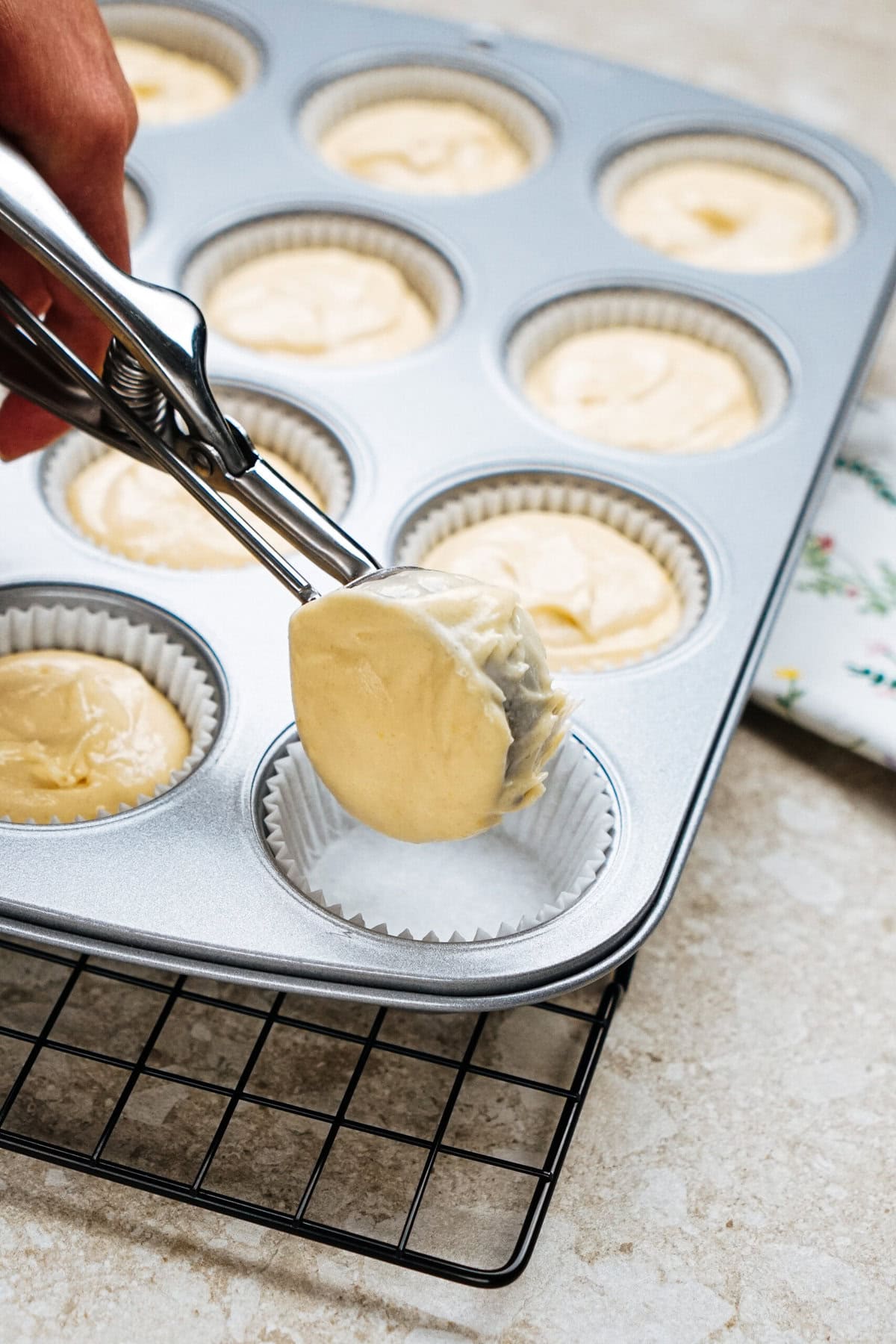 Hand using a scoop to fill a muffin tray with batter, placed on a wire rack.