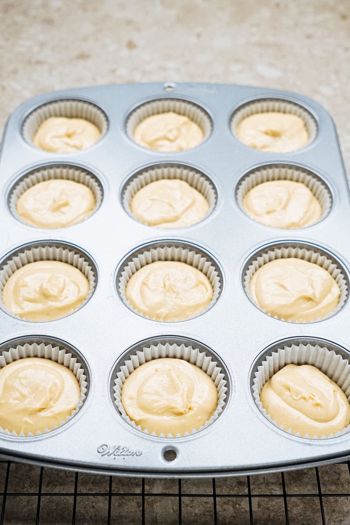 Muffin tin with twelve paper liners filled with uncooked vanilla batter, placed on a wire rack.