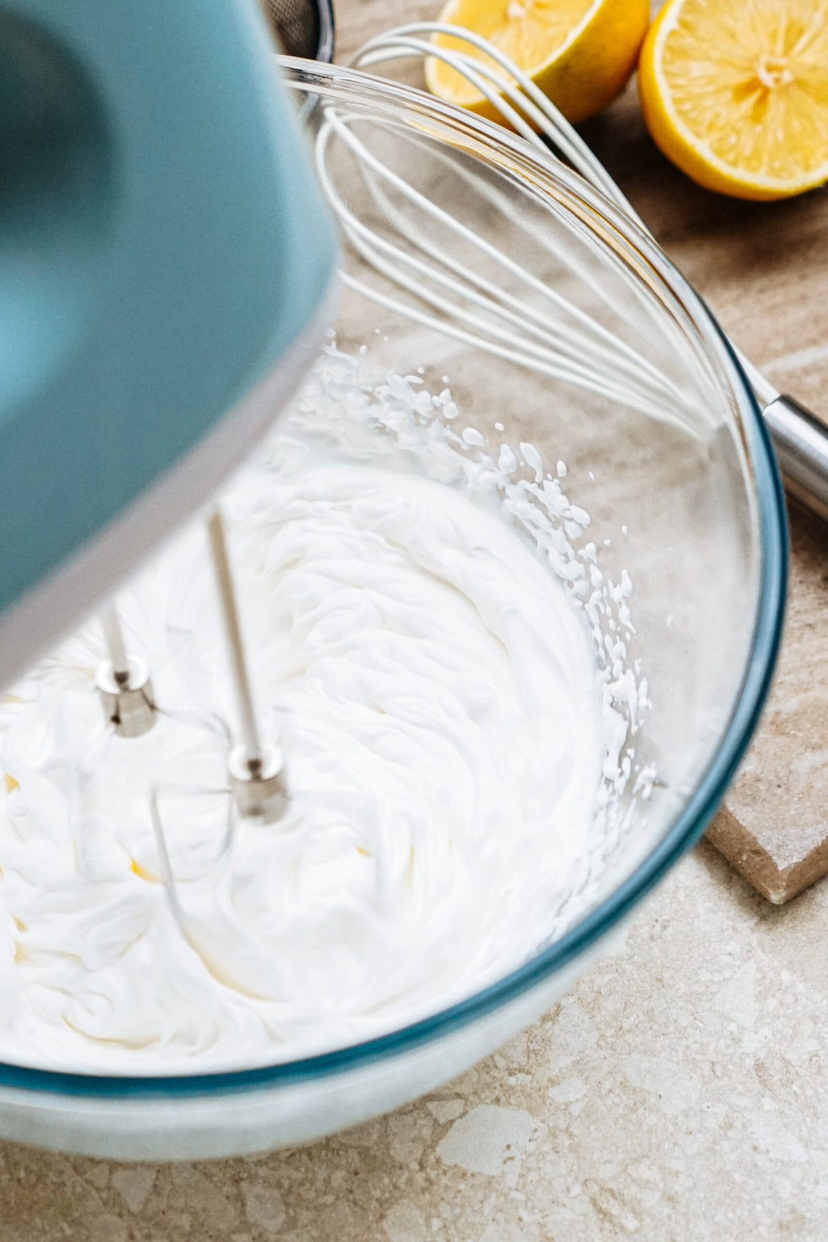 An electric mixer is whipping cream in a glass bowl, with cut lemons and a whisk in the background on a marble countertop.