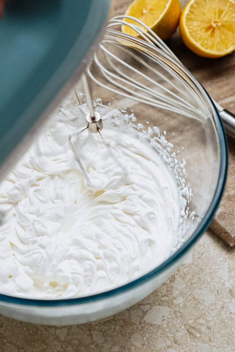 Electric mixer whipping cream in a glass bowl, with halved lemons and a whisk nearby on a countertop.