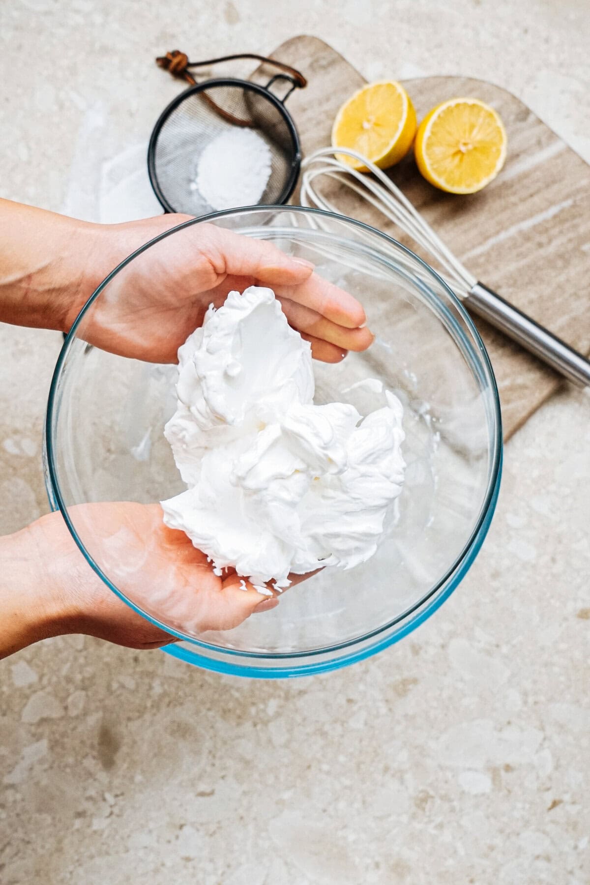 Two hands holding a glass bowl of whipped cream over a countertop with a whisk, halved lemons, and a sifter in the background.