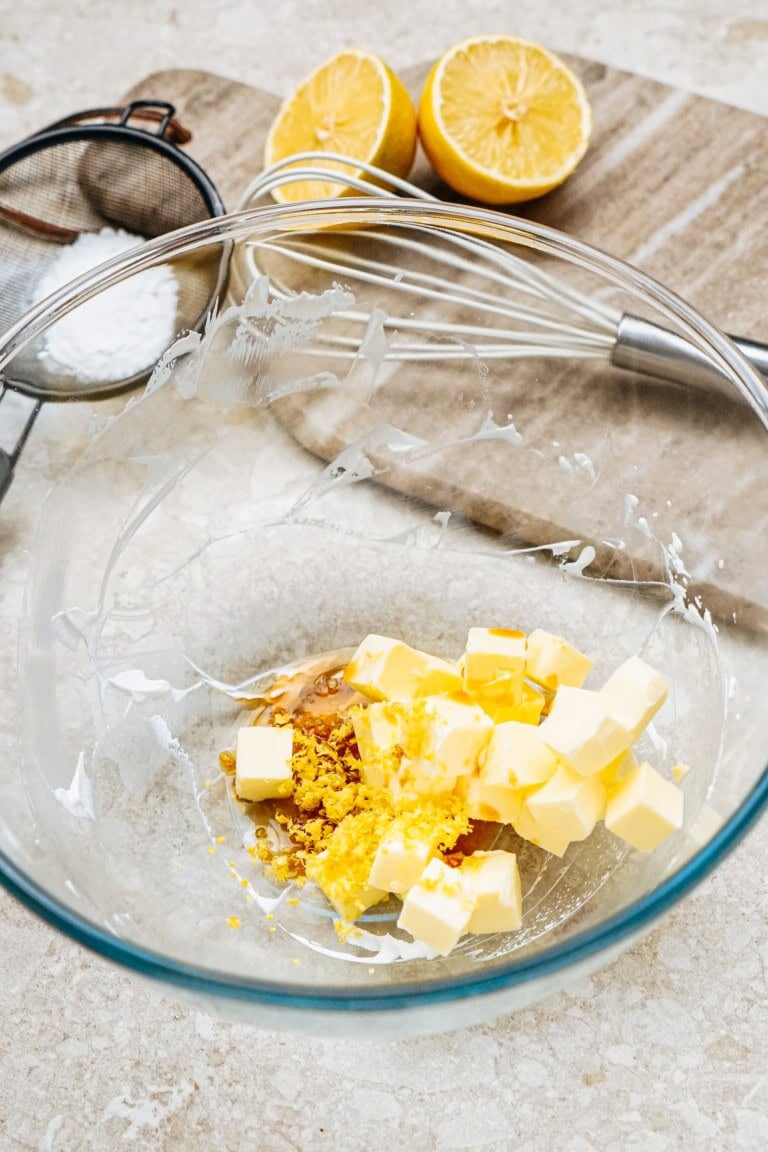 Cubed butter and lemon zest in a glass bowl, with a whisk, sifter, powdered sugar, and halved lemons on a cutting board in the background.