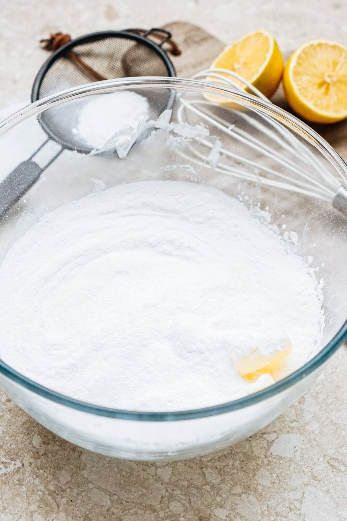 A glass bowl of flour with a small butter piece on top, next to a whisk and sieve. Two halved lemons are on the table.