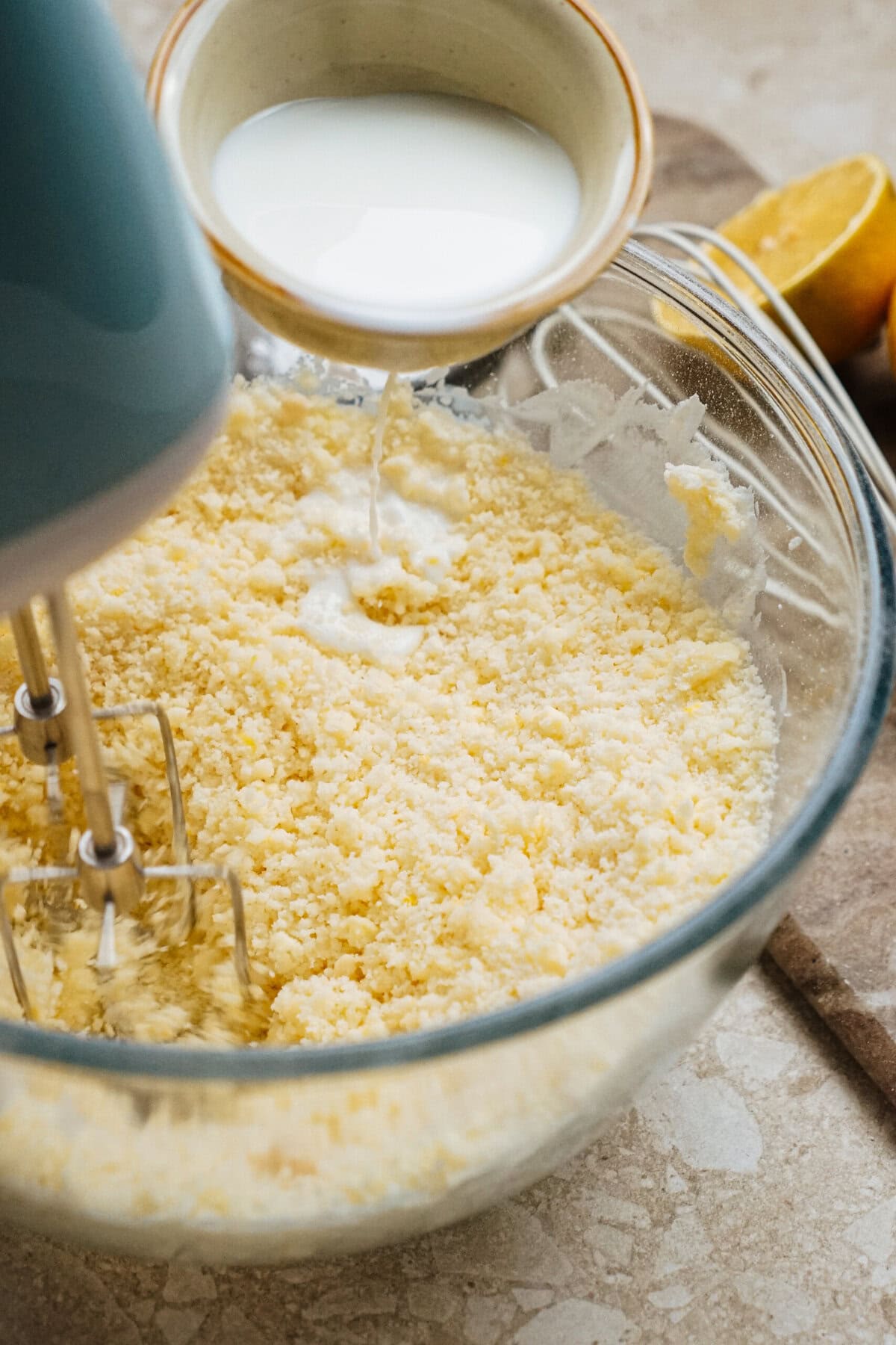 Mixer blending a mixture in a glass bowl as milk is being poured in. Nearby are a half lemon and a cloth.