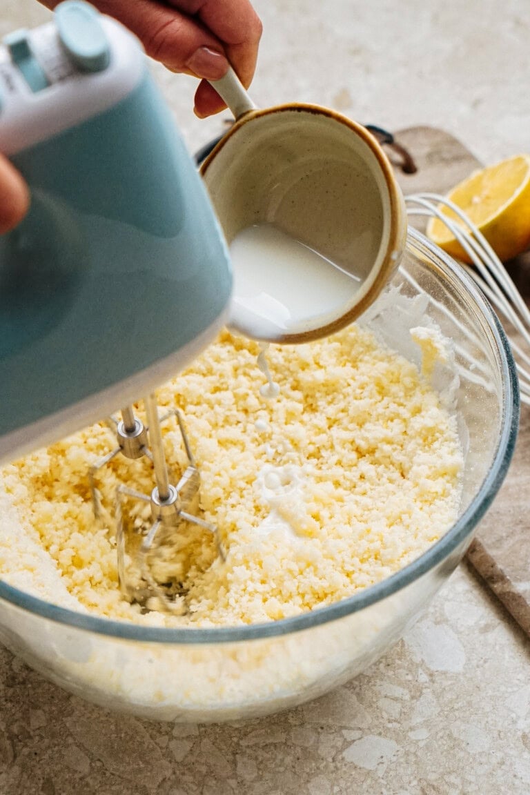 Hands pouring milk into a bowl with electric beaters mixing dough, with a lemon and a spoon nearby.