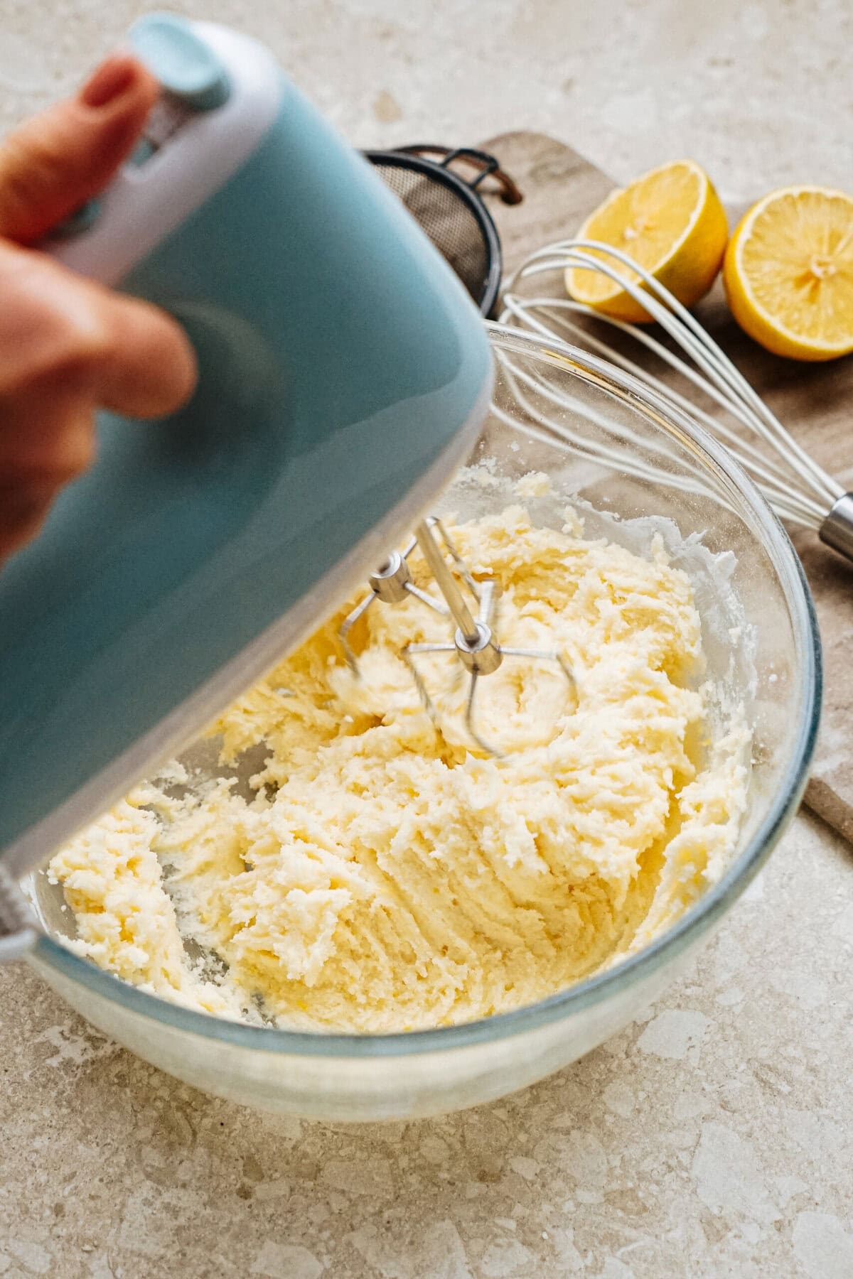 A person uses a hand mixer to blend ingredients in a glass bowl. Two halved lemons and a whisk are on the countertop nearby.