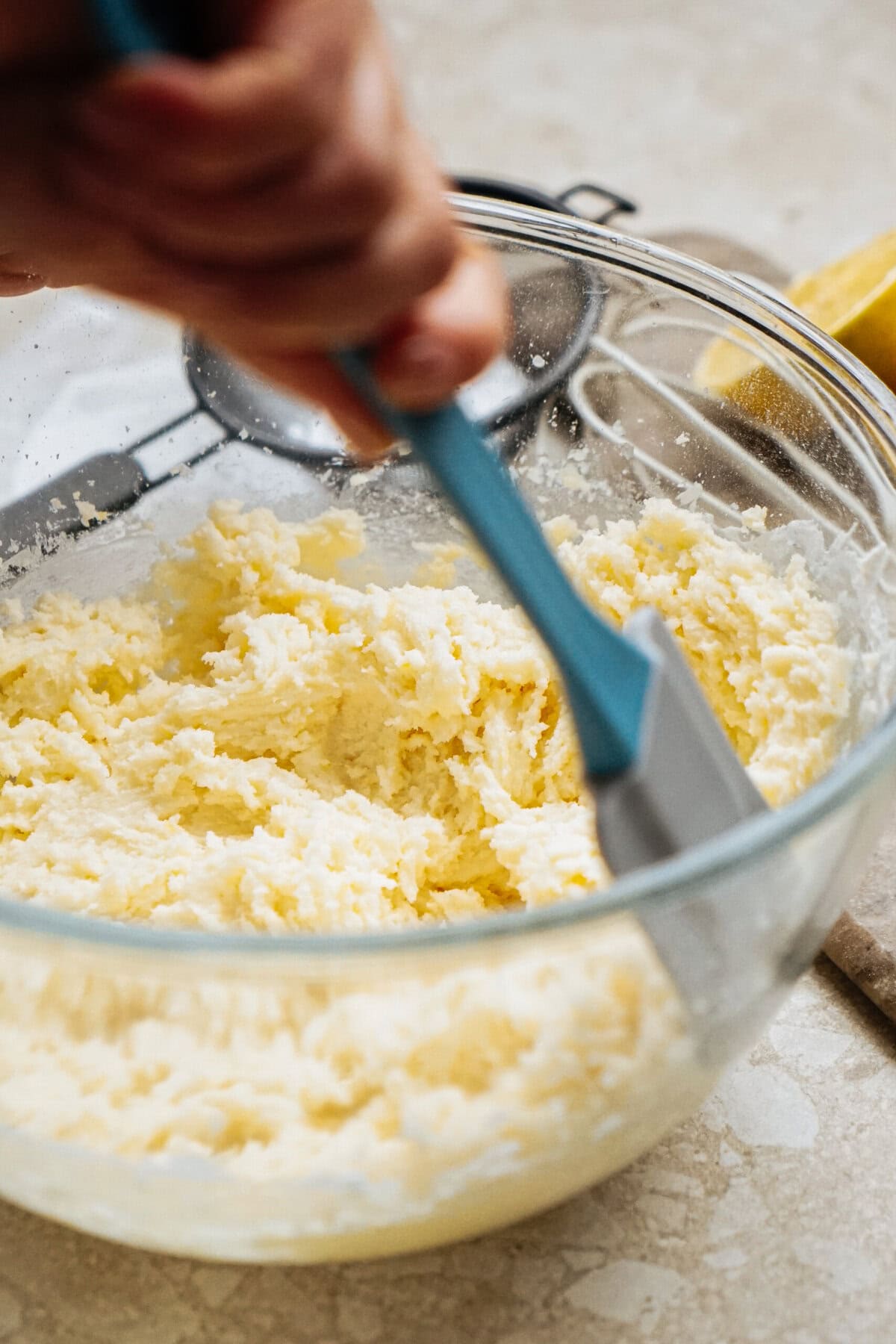 Person mixing butter and sugar in a glass bowl with a blue spatula.