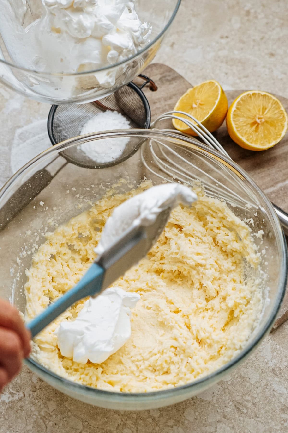 A mixing bowl with yellow batter and a spatula adding whipped cream. Two halved lemons and an empty glass bowl are in the background.